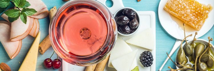 Summer wine snack set. Glass of rose, meat, cheese, olives, honey, bread sticks, nuts, capers and berries with white ceramic board in center, blue wooden background, top view